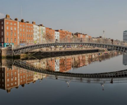 A metal pedestrian bridge spans a calm river with brick buildings and their reflections visible on either side under a clear sky.