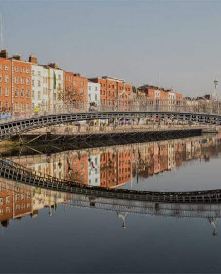 A metal pedestrian bridge spans a calm river with brick buildings and their reflections visible on either side under a clear sky.