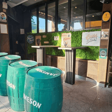 Bar entrance with green Jameson barrels used as tables, high-top tables, vintage beer signs on a grass-covered wall, and a staircase leading inside.
