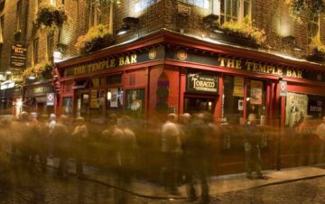 A crowd of people gathers outside The Temple Bar pub at night, with blurred motion suggesting activity. The red facade and hanging plants are illuminated by streetlights.