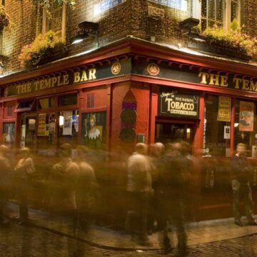 A crowd of people gathers outside The Temple Bar pub at night, with blurred motion suggesting activity. The red facade and hanging plants are illuminated by streetlights.