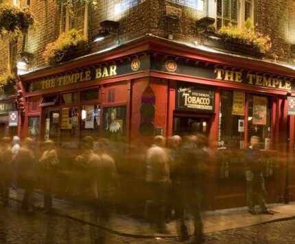 A crowd of people gathers outside The Temple Bar pub at night, with blurred motion suggesting activity. The red facade and hanging plants are illuminated by streetlights.