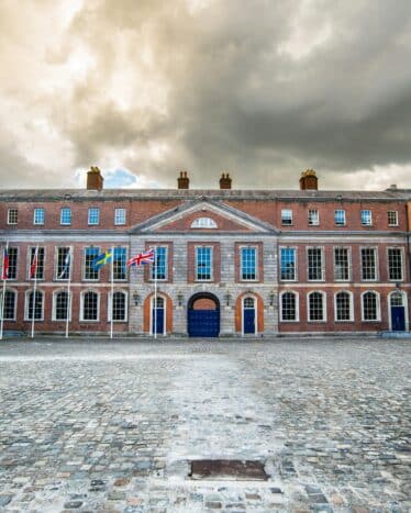 A large cobblestone courtyard surrounded by a historic brick building with many windows and a row of international flags near the entrance under a cloudy sky.