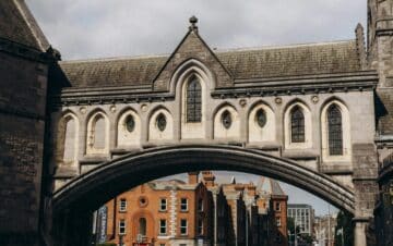 A stone arch bridge connects two historic buildings over a city street with pedestrians and a cyclist below.