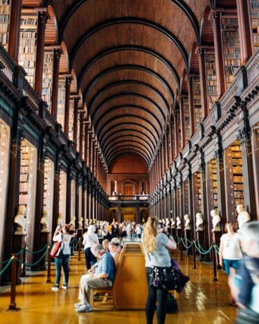 People walk and sit in a large, historic library hall with high arched ceilings, tall bookshelves, and numerous statues lining the central aisle.