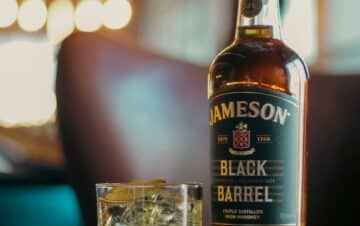 A bottle of Jameson Black Barrel whiskey and a glass filled with whiskey and ice sit on a wooden table in a warmly lit room.