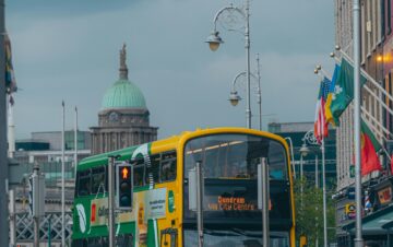 People walk near a yellow double-decker bus at a city street intersection, with historic buildings and a domed structure visible in the background under a cloudy sky.