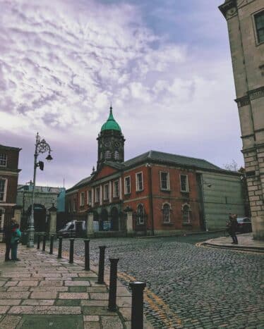 A cobblestone street with bollards leads to a historic red brick building with a green-domed clock tower under a partly cloudy sky.